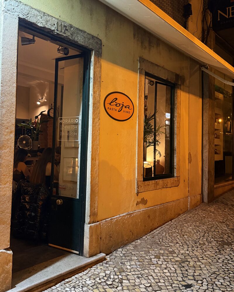 A fachada à noite: a porta verde aberta para a sala iluminada, a placa oval na parede creme e a calçada à frente