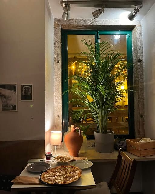 A sala à noite: mesa para dois com uma pizza, uma palmeira em vaso e a janela de caixilho verde