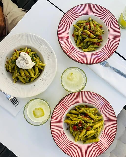 Três pratos de penne ao pesto, um com burrata, numa mesa branca